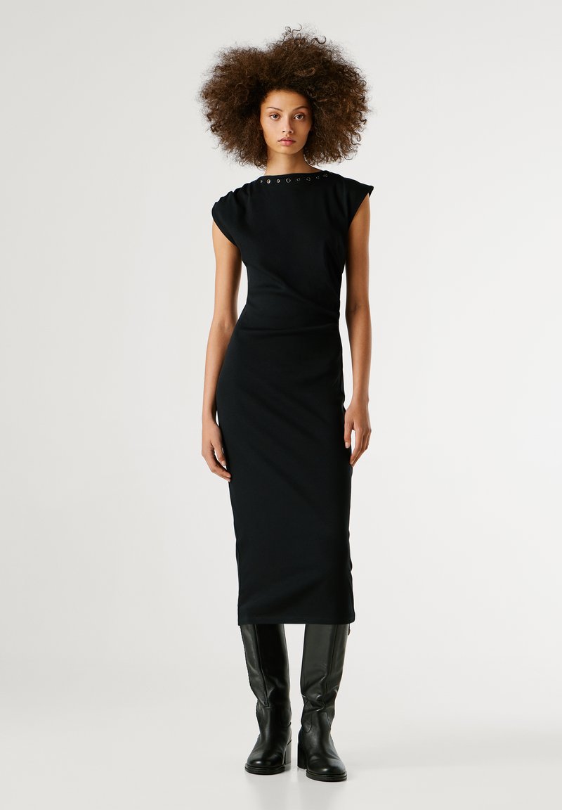 Abito nero midi con forma aderente, maniche corte, tessuto testurizzato e accenti metallici decorativi attorno al colletto; abbinato a stivali neri sopra il ginocchio.