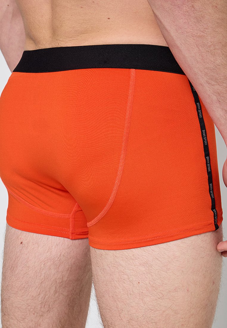 Orange Boxershorts mit einem schwarzen Elastikbund, die eine enge Passform, flache Nähte und eine seitliche Akzentuierung mit dem Markennamen aufweisen.