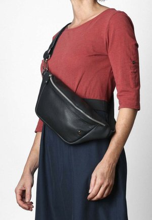 Borsa a tracolla in pelle nera con zip diagonale e texture liscia. Indossata sopra un top rosso e una gonna scura, con tracolla regolabile e dettagli in metallo.