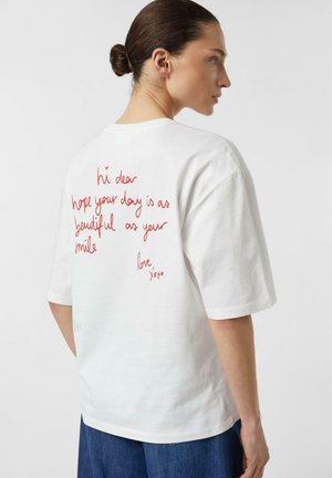 Kobieta z brązowymi włosami upiętymi w kok, ubrana w białą koszulkę z czerwoną, odręcznie napisaną treścią na plecach, stojąca na jednolitym jasnym tle.