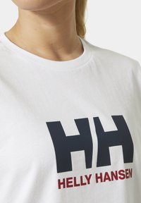 Primer plano de una camiseta blanca de cuello redondo con grandes letras negras "HH" y texto rojo "HELLY HANSEN" en el pecho.