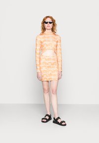 Vestido de manga longa cortado em laranja com um padrão desenhado. Combinado com sandálias pretas e óculos de sol. Textura suave.