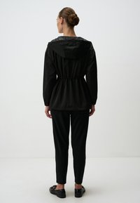 Zwarte lichtgewicht jas met een koord in de taille, capuchon en lange mouwen; gecombineerd met zwarte slim-fit broeken en instapschoenen.