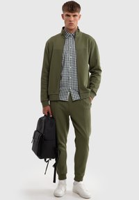 Olivenfarvet hættejakke med lynlås og matchende joggers, med en hvid- og grønternet skjorte under. Modellen holder en sort rygsæk.