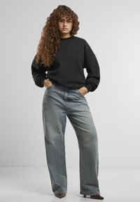 Felpa nera oversize con scollatura tonda abbinata a jeans a gamba larga blu chiaro. Il modello sta in piedi con le mani nelle tasche, con capelli mossi.