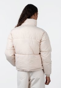 Puffete, kort jakke i lys rosa, med høy krage, lange ermer og elastiske mansjetter. Glatt, blank tekstur med sydde paneler.