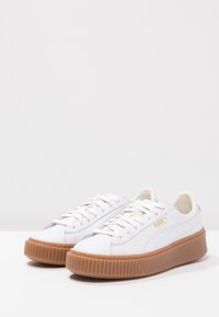 Zapatillas de piel sintética blanca con suela de goma texturizada, que cuentan con un diseño de cordones y un sutil acabado dorado en el lateral.