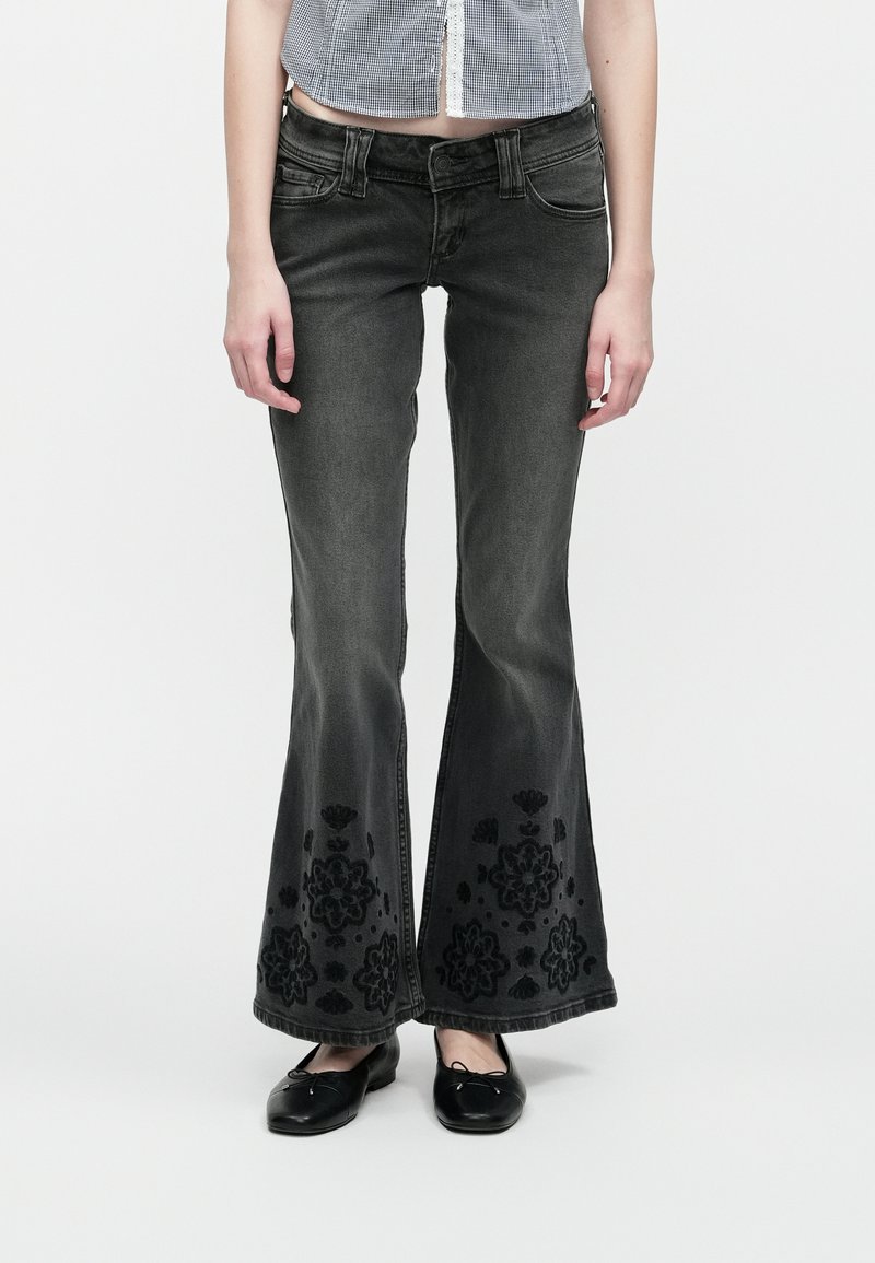 Sieviete valkā tumšus platās kājas džinsus ar melniem ziedu izšuvumiem pie apakšmalas un melnām balerīnu kurpēm, stāvot pret vienkāršu fonu.