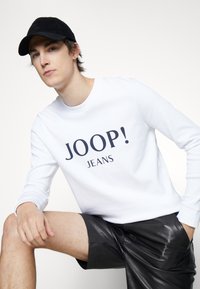 Felpa bianca con la scritta "JOOP! JEANS" stampata in blu navy, abbinata a pantaloni corti di pelle nera. La modella indossa un cappellino nero, posando in modo informale.