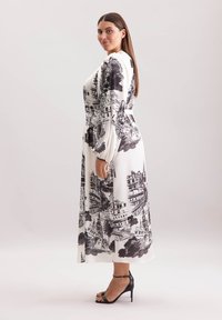 Langes, weißes Kleid mit einem schwarzen Stadtbild-Print. Fließender Stoff mit langen Ärmeln und tailliertem Schnitt, kombiniert mit schwarzen High-Heels.