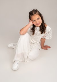 Weiße plissierte Hose, Spitzenoberteil mit Blumenmuster und weiße Sneaker. Sitzpose mit Händen unter dem Kinn, lange Haare, neutraler Hintergrund.