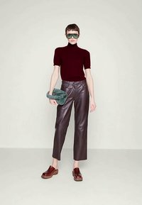 Μπορντό ζιβάγκο, καφέ δερμάτινα παντελόνια σε φαρδιά γραμμή, πράσινη γούνα τσάντα, και καφέ κλόγκ. Το μοντέλο φορά πράσινα γυαλιά ηλίου. Ουδέτερο φόντο.