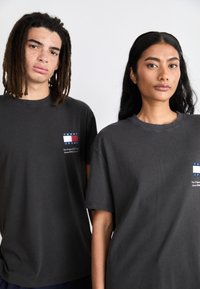 Camisetas de algodón gris con mangas cortas que presentan un logo con rayas rojas, blancas y azules en el lado izquierdo del pecho. Diseño simple, ajuste unisex.