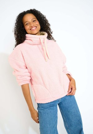 Pullover in pile rosa con collo alto, dettagli con cordino e superficie testurizzata; abbinato a jeans a gamba larga blu chiaro.