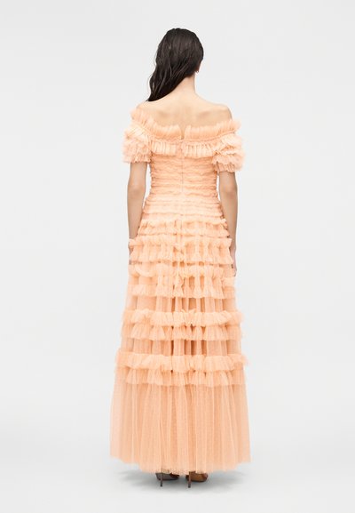 Selja tagant vaadatav naine kannab pikka virsikuvärvi off-shoulder kleiti, millel on kihilised volangid ja kroogitud ülakehaosa.