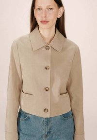 Donna che indossa una giacca beige in suede con bottoni, colletto e due tasche frontali, abbinata a jeans blu in denim a vita alta.