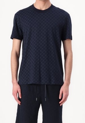 Mężczyzna ubrany w granatową koszulę z krótkim rękawem i dopasowane granatowe spodnie z subtelnym geometrycznym wzorem oraz elastycznym paskiem w talii.