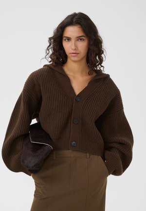 Donna con capelli scuri e ricci che indossa un cardigan marrone a coste con grandi bottoni e tiene una pochette scura in pelliccia.