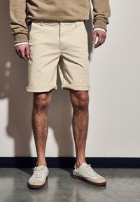 Pantalones cortos de algodón beige con dobladillos enrollados, con bolsillos frontales. Combinados con zapatillas blancas que presentan acentos de cuero y gamuza.