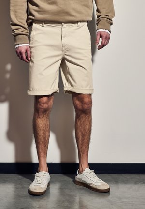 Beige bomulds shorts med opfoldede manchetter, med frontlommer. Parret med hvide sneakers, der viser læder- og ruskindselementer.