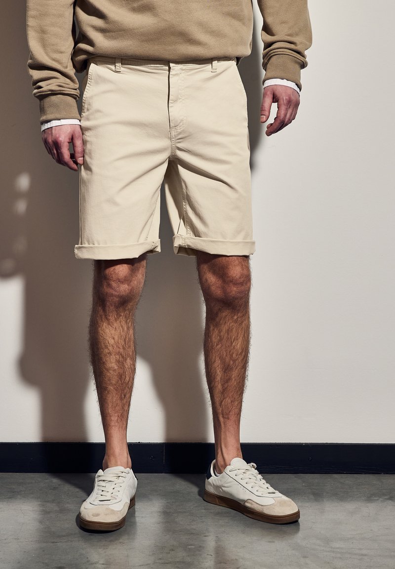 Pantalones cortos de algodón beige con dobladillos enrollados, con bolsillos frontales. Combinados con zapatillas blancas que presentan acentos de cuero y gamuza.