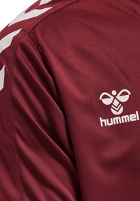 Bordeauxrode sportjas schouderdeel met wit hummel bij-logo en chevronstrepen op de mouw.