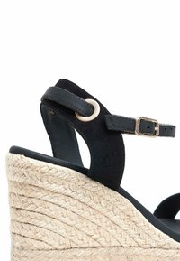 Zwarte suède wedge sandaal met jute platform, verstelbare enkelband en goudkleurige hardwaredetails. Gelaagd geweven ontwerp op de wedge.