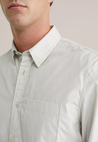 Šviesiai pilkas mygtukinis marškinis, pagamintas iš lygios medvilnės, su klasikiniu apykakle, kišene kairiajame krūtinės plote ir subtilių siuvimo detalių.
