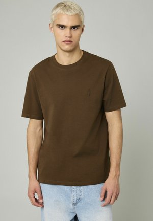 T-shirt en coton marron avec col rond et manches courtes. Comprend un subtil détail de broderie sur la poitrine. Assorti à un denim bleu clair.