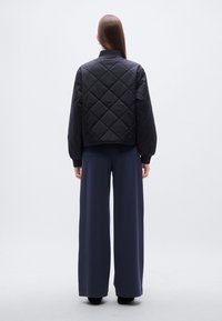 Μαύρο μπουφάν bomber με καπιτονέ σχέδιο, κοντό μήκος, ριμπ μανσέτες και ψηλό γιακά, συνδυασμένο με μπλε σκούρο παντελόνι με φαρδιά μπατζάκια. Λείο υλικό.