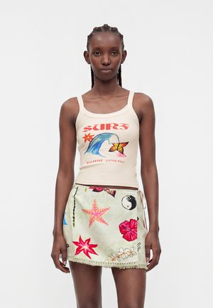 Femme aux cheveux tressés portant un débardeur crème avec un motif surf et une jupe portefeuille fleurie avec des motifs étoiles de mer et yin-yang.