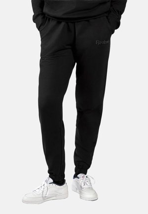 Zwarte joggerbroek met elastische boorden, zijzakken en een klein logo op het dijbeen, gedragen met witte sneakers en handen in de zakken.