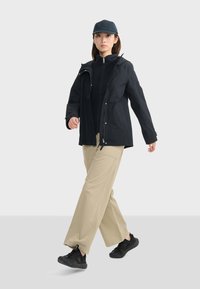Veste imperméable noire avec capuche, haut noir zippé en dessous, pantalon large beige, et baskets noires, le tout porté avec une casquette bleu marine.