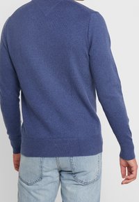 Homme portant un pull en tricot bleu et un jean bleu clair, vu de dos sur un fond blanc uni.