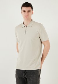 Herren Poloshirt in hellbeige mit kurzem Reißverschluss, Kragen, kurzen Ärmeln und einem glatten, dehnbaren Stoff. Kombiniert mit dunklen Jeans.