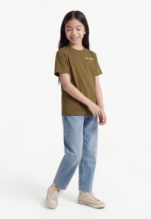 Κορίτσι που χαμογελά, με φανέλα Off-White σε ελαιί πράσινο, ανοιχτόχρωμα μπλε τζιν, και μπεζ ψηλά αθλητικά παπούτσια, στέκεται μπροστά σε λευκό φόντο.