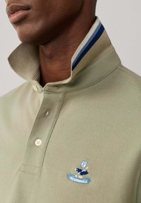 Gros plan d'une personne portant un polo beige à col relevé avec des rayures bleues et le logo oiseau Façonnable sur la poitrine.