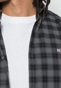 Grått rutigt skjorta med knapp-up lager på en vit crew neck t-shirt, med subtil textur och en liten varumärkeslogotyp på bröstet.