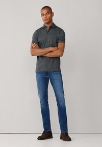 Šedé polo tričko s krátkým rukávem, ve spojení s modrými džíny. Model stojí s pažemi zkříženými, na nohách má hnědé boty. Hladká látka, přiléhavý střih.