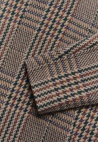 Tkanina wełniana w wzór houndstooth w kolorach beżowym, bordowym, zielonym i granatowym. Tekstura wydaje się miękka i ciężka. Krawędzie są obszyte.