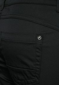 Gros plan d'une poche arrière en tissu noir sur un pantalon avec des coutures visibles et une petite rivet métallique dans le coin supérieur.