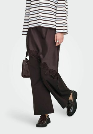 Personne portant un pantalon noir large avec bordure en dentelle, chemise à manches longues rayée, mocassins noirs vernis, tenant un petit sac à main noir.