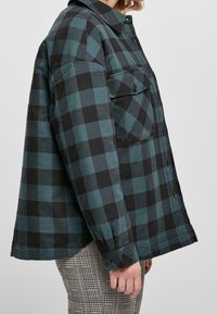 Camisa de franela a cuadros verdes y negros con cuello abotonado, dos bolsillos en el pecho y mangas largas. Textura suave y ajuste relajado.