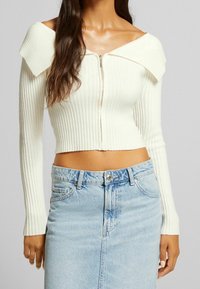 Biały prążkowany krótki sweter z odkrytym kołnierzem, z przodu zapinany na zamek, z długimi rękawami, w połączeniu z jasnoniebieską dżinsową spódnicą ołówkową.