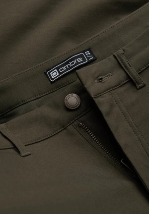 Pantalon vert olive avec une texture lisse, doté d'une fermeture éclair texturée, d'un bouton de marque et d'une étiquette de marque près de la taille.