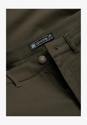 Pantaloni verde oliva con una texture liscia, dotati di una cerniera testurizzata, un bottone marchiato e un'etichetta con il marchio vicino alla vita.