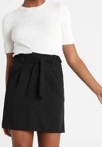 Top bianco a maniche corte e costine con scollatura rotonda abbinato a una gonna nera a vita alta con cintura da annodare e parte frontale plissettata.