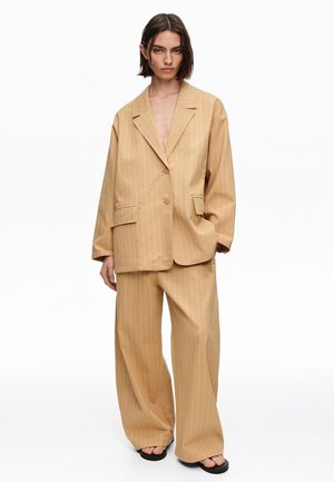 Persona con un traje de raya diplomática beige de gran tamaño con pantalones de pierna ancha y sandalias negras, de pie con una mano en el bolsillo.