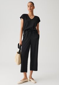 Zwart jumpsuit met een V-hals, korte mouwen, een trekkoord in de taille en wijde benen. Gecombineerd met beige flats en een textuur, ronde strooien tas.