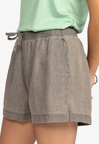 Šedé šortky s vysokým pasem, pružným pasem a šňůrkou, nošené s lehce zeleným topem zašněrovaným na boku, boční pohled na dolní část těla osoby.